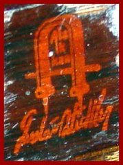 Close-up of the Gustav Stickley red decal signature on the back of the rear stretcher. "Gustav Stickley" in script below the craftsman joiners mark surrounding the words: "Als Ik Kan", translated: ....."as I can" or "to the best of my ability"